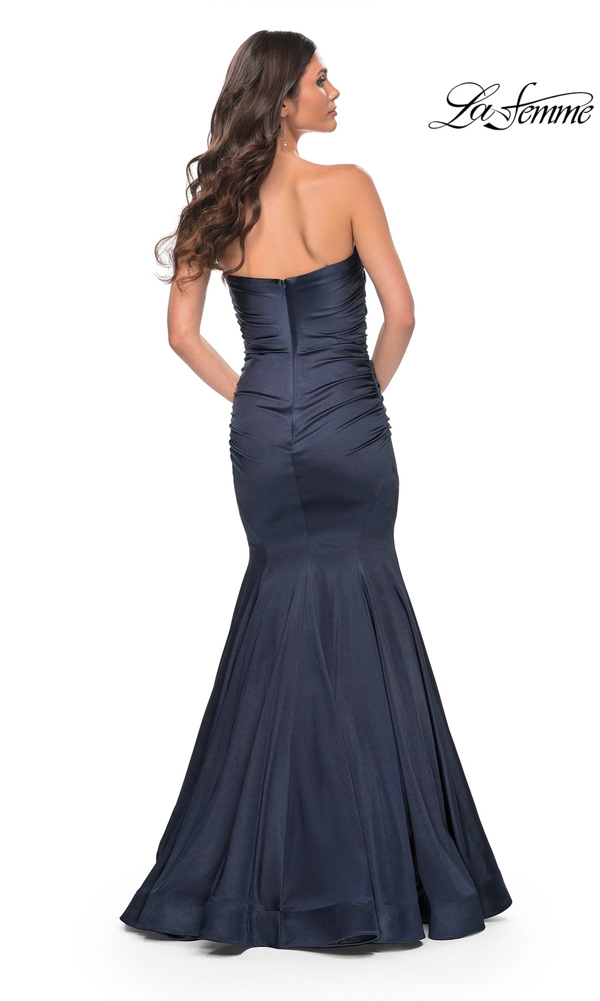 A woman with long brown hair wears the La Femme Strapless Long Mermaid Prom Dress 31980, a navy blue, floor-length gown with subtle ruching and a fitted bodice, shown from the back.